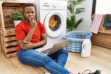 Young african man doing laundry and using computer looking confident at the camera smiling with crossed arms and hand raised on chin. thinking positive.