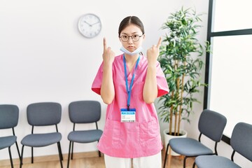 Young asian nurse woman at medical waiting room pointing up looking sad and upset, indicating direction with fingers, unhappy and depressed.