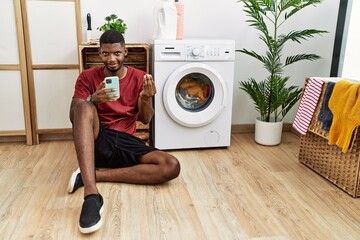 Young african american man using smartphone waiting for washing machine doing money gesture with...
