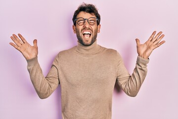 Handsome man with beard wearing turtleneck sweater and glasses celebrating mad and crazy for...