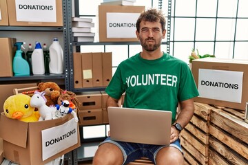 Young handsome man wearing volunteer t shirt using laptop puffing cheeks with funny face. mouth...