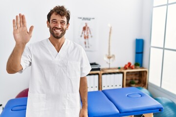 Young handsome physiotherapist man working at pain recovery clinic waiving saying hello happy and smiling, friendly welcome gesture