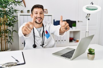 Obraz premium Young doctor working at the clinic using computer laptop approving doing positive gesture with hand, thumbs up smiling and happy for success. winner gesture.