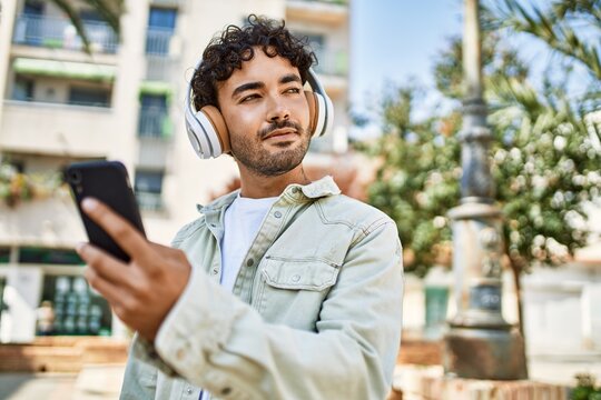 Handsome hispanic man with beard smiling happy outdoors on a sunny day wearing headphones listening to music with smartphone