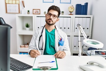 Young man with beard wearing doctor uniform and stethoscope at the clinic relaxed with serious expression on face. simple and natural looking at the camera.