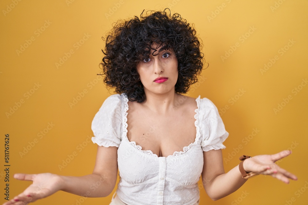 Sticker young brunette woman with curly hair standing over yellow background clueless and confused with open