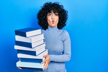 Young middle east woman holding a pile of books making fish face with mouth and squinting eyes, crazy and comical.