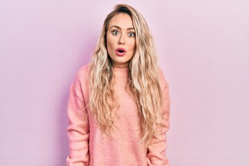 Beautiful young blonde woman wearing pink sweater scared and amazed with open mouth for surprise,...