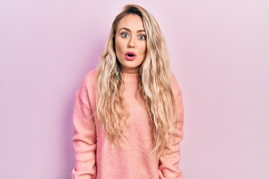 Beautiful young blonde woman wearing pink sweater scared and amazed with open mouth for surprise, disbelief face