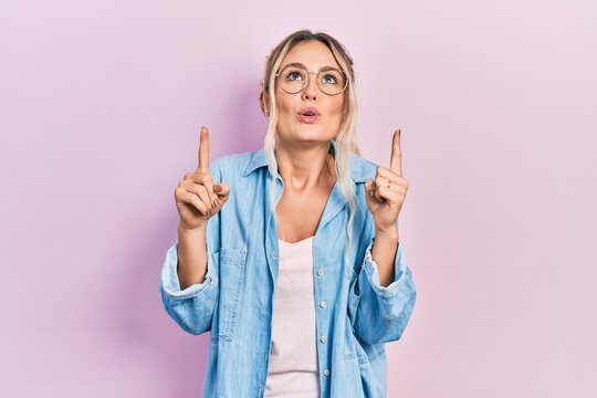 Beautiful young blonde woman wearing casual clothes and glasses amazed and surprised looking up and pointing with fingers and raised arms.