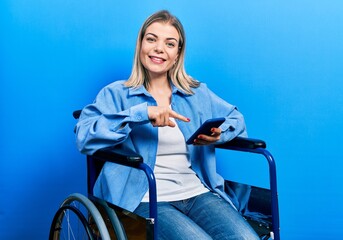 Beautiful caucasian woman sitting on wheelchair using smartphone smiling happy pointing with hand...