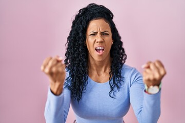 Middle age hispanic woman standing over pink background angry and mad raising fists frustrated and...