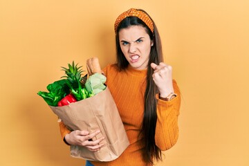 Young brunette teenager holding paper bag with groceries smiling cheerful pointing with hand and finger up to the side