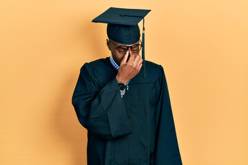 Young african american man wearing graduation cap and ceremony robe tired rubbing nose and eyes...