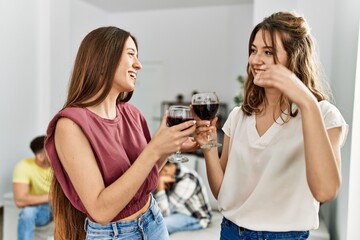 Group of young friends smiling happy sitting on the sofa. Two women toasting with glass of red wine at home.