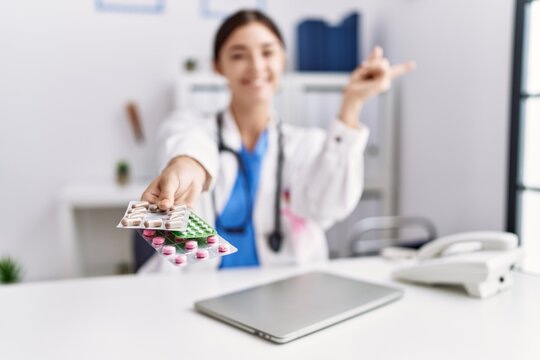 Young hispanic doctor woman wearing doctor uniform holding prescription pills at clinic smiling happy pointing with hand and finger to the side