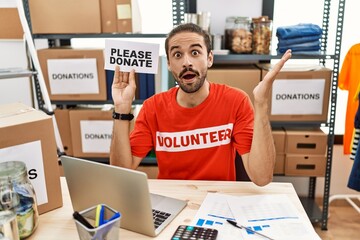 Young hispanic man wearing volunteer t shirt holding please donate banner celebrating achievement with happy smile and winner expression with raised hand