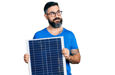Hispanic man with beard holding photovoltaic solar panel smiling looking to the side and staring...