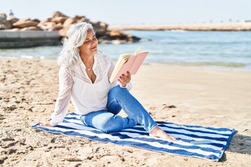 Middle age woman reading book sitting on towel at seaside