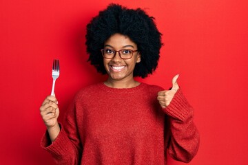 Young african american woman holding one silver fork smiling happy and positive, thumb up doing...