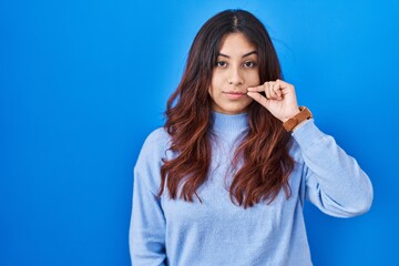 Hispanic young woman standing over blue background mouth and lips shut as zip with fingers. secret...