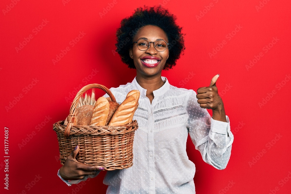 Wall mural young african american woman holding wicker basket with bread smiling happy and positive, thumb up d