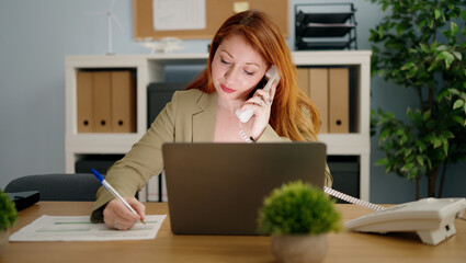 Young redhead woman business worker using laptop talking on telephone at office