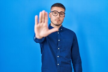Young hispanic man wearing glasses over blue background doing stop sing with palm of the hand....