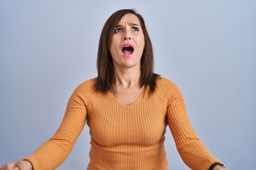 Middle age brunette woman standing wearing orange sweater angry and mad screaming frustrated and...