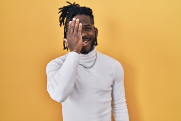 African man with dreadlocks wearing turtleneck sweater over yellow background covering one eye with hand, confident smile on face and surprise emotion.