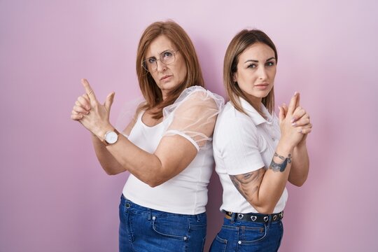 Hispanic mother and daughter wearing casual white t shirt over pink background holding symbolic gun with hand gesture, playing killing shooting weapons, angry face