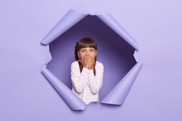 Horizontal shot of shocked scared little girl with braids looking through breakthrough of purple...