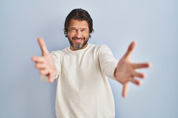 Fototapeta premium Handsome middle age man wearing casual sweater over blue background looking at the camera smiling with open arms for hug. cheerful expression embracing happiness.