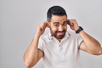 Young hispanic man with beard wearing casual clothes over white background smiling pulling ears with fingers, funny gesture. audition problem