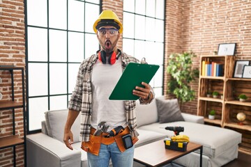 Young hispanic man with beard working at home renovation scared and amazed with open mouth for...