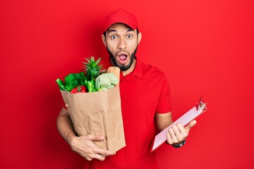 Hispanic man with beard wearing courier uniform with groceries from supermarket and clipboard afraid and shocked with surprise and amazed expression, fear and excited face.