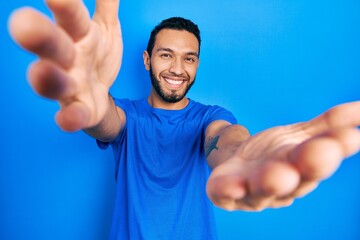 Hispanic man with beard wearing casual blue t shirt looking at the camera smiling with open arms for hug. cheerful expression embracing happiness.