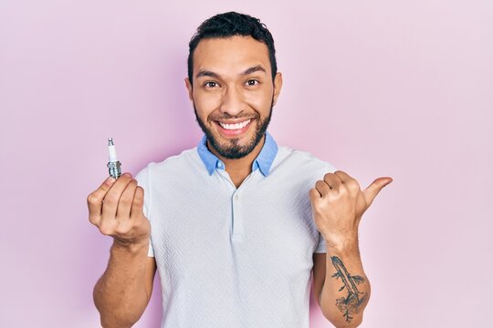 Hispanic man with beard holding spark plug pointing thumb up to the side smiling happy with open mouth