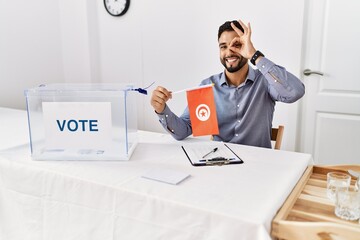 Young handsome man with beard at political campaign election holding tunisia flag smiling happy doing ok sign with hand on eye looking through fingers