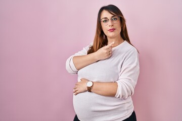 Fototapeta premium Pregnant woman standing over pink background pointing with hand finger to the side showing advertisement, serious and calm face
