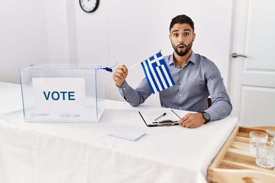Young handsome man with beard at political campaign election holding greece flag scared and amazed with open mouth for surprise, disbelief face