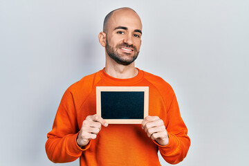 Young bald man holding blackboard looking positive and happy standing and smiling with a confident smile showing teeth