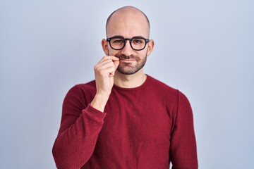 Young bald man with beard standing over white background wearing glasses mouth and lips shut as zip...