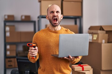 Young bald man with beard working at small business ecommerce with laptop angry and mad screaming frustrated and furious, shouting with anger looking up.