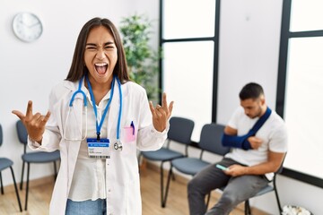 Young asian doctor woman at waiting room with a man with a broken arm shouting with crazy expression doing rock symbol with hands up. music star. heavy concept.