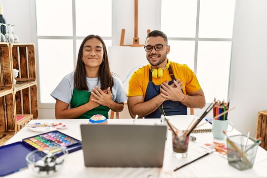 Young hispanic couple using laptop sitting on the table at art studio smiling with hands on chest with closed eyes and grateful gesture on face. health concept.