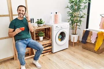 Middle age hispanic man doing laundry talking on the smartphone and drinking coffee at home.
