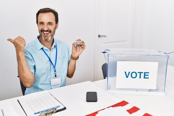 Middle age man with beard sitting by ballot holding i vote badge smiling with happy face looking and pointing to the side with thumb up.