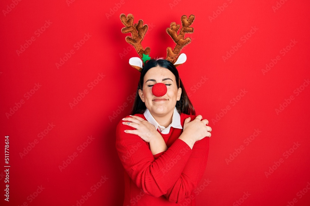 Sticker young hispanic woman wearing deer christmas hat and red nose hugging oneself happy and positive, smi