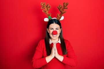 Young hispanic woman wearing deer christmas hat and red nose praying with hands together asking for forgiveness smiling confident.
